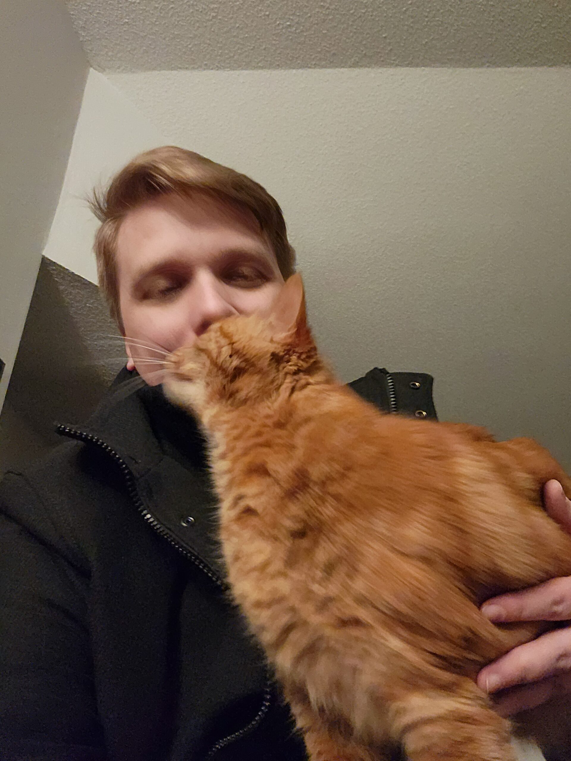 A blurry selfie of a man in a black coat cuddling an orange tabby cat.